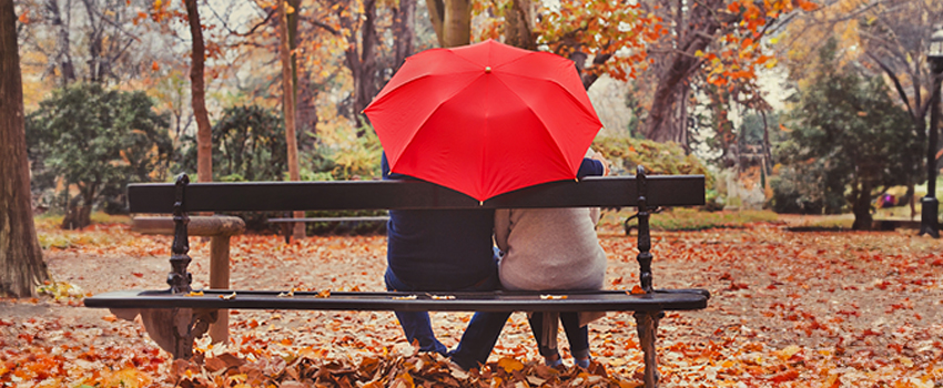 A couple sitting on a park bench in Autumn. They are facing away from the viewer, and are sharing a bright red umbrella.